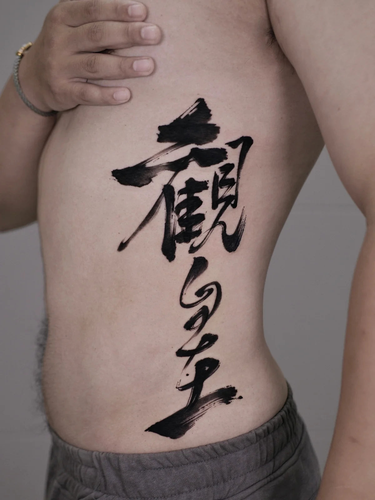 Vertical black Chinese brush calligraphy tattoo on the side of the torso, with broad upper strokes and a sweeping lower stroke